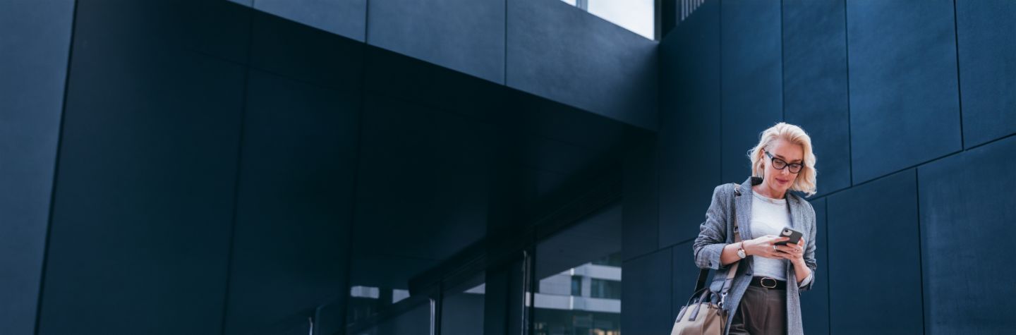 Une femme d'affaires blonde en blazer gris se tient devant un bâtiment moderne, regardant son smartphone et souriant.