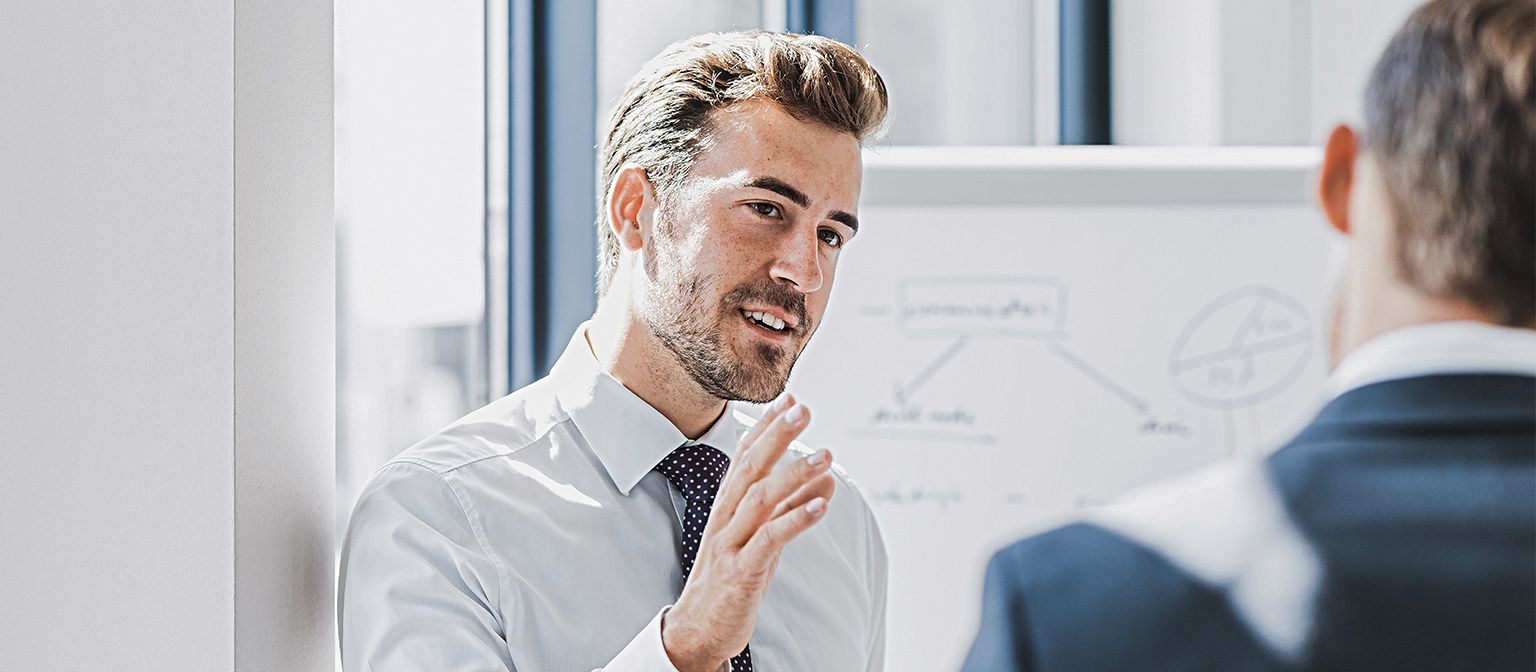 On voit un homme en tenue professionnelle expliquer quelque chose avec une expression amicale et engagée. Face à lui se tient une autre personne qui l’écoute attentivement. On aperçoit en arrière-plan un tableau blanc avec un croquis.