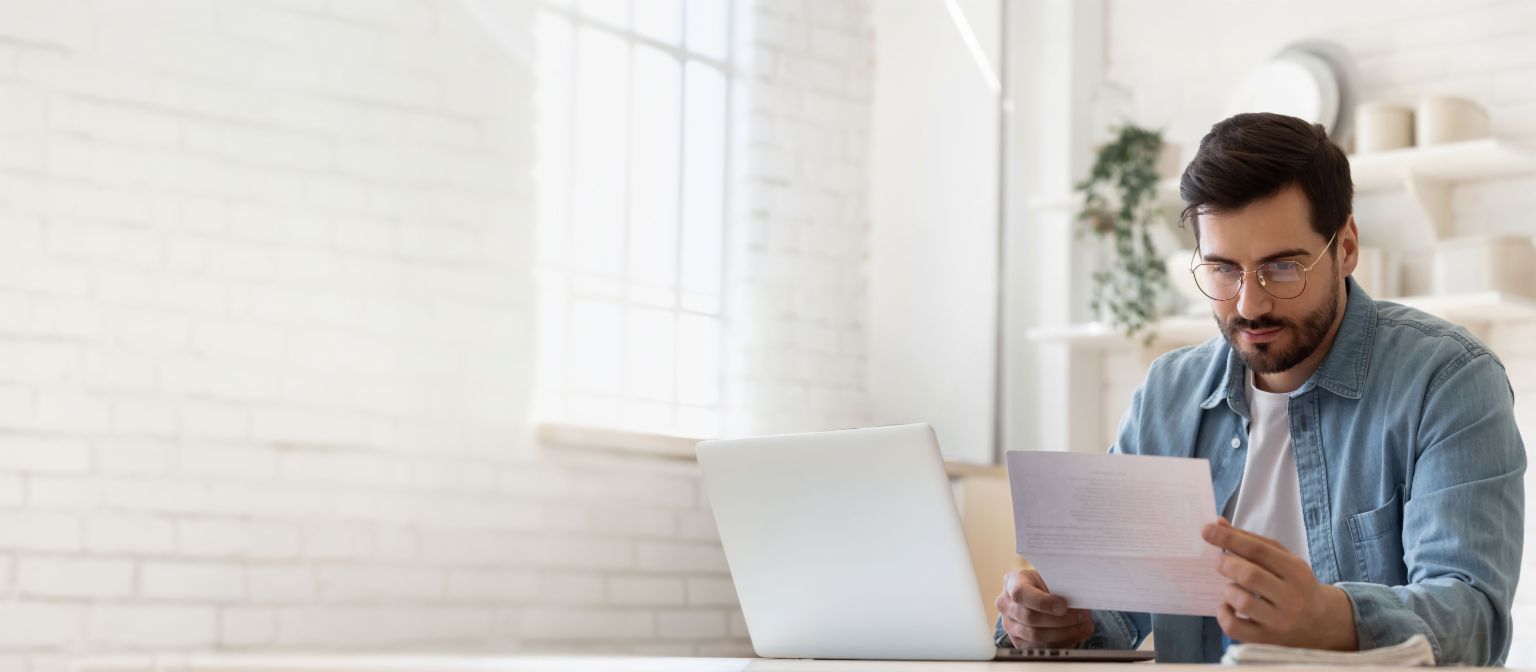 Homme portant des lunettes travaillant sur ordinateur portable tout en examinant un document de règlement de dettes dans une maison lumineuse