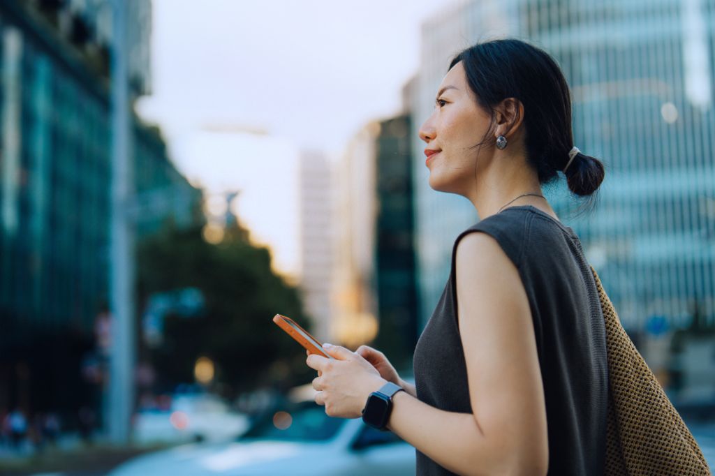 Une jeune femme se tient dehors dans un environnement urbain, tenant son smartphone et souriant en regardant devant elle. Des bâtiments modernes et des arbres sont visibles en arrière-plan.