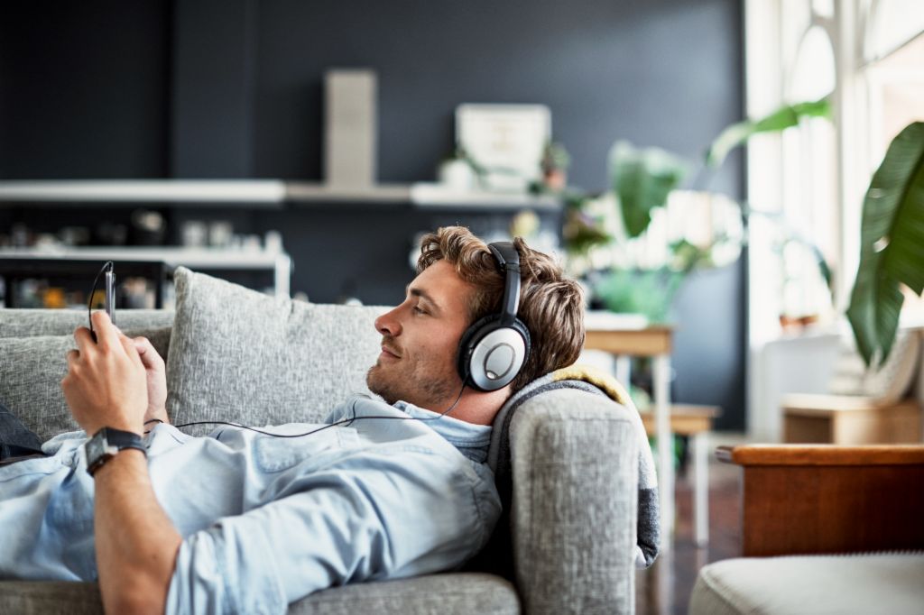Un jeune homme est allongé sur un canapé dans son salon, portant un casque et regardant son smartphone en souriant, tout en écoutant de la musique ou un podcast.