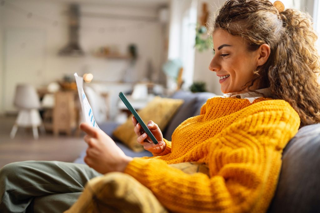 Une jeune femme souriante aux cheveux bouclés, vêtue d'un pull jaune confortable, est assise sur un canapé dans son salon et regarde son smartphone tout en tenant un document papier.