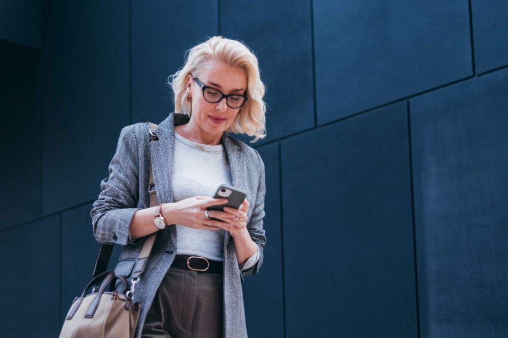 Une femme d'affaires blonde en blazer gris se tient devant un bâtiment moderne, regardant son smartphone et souriant.