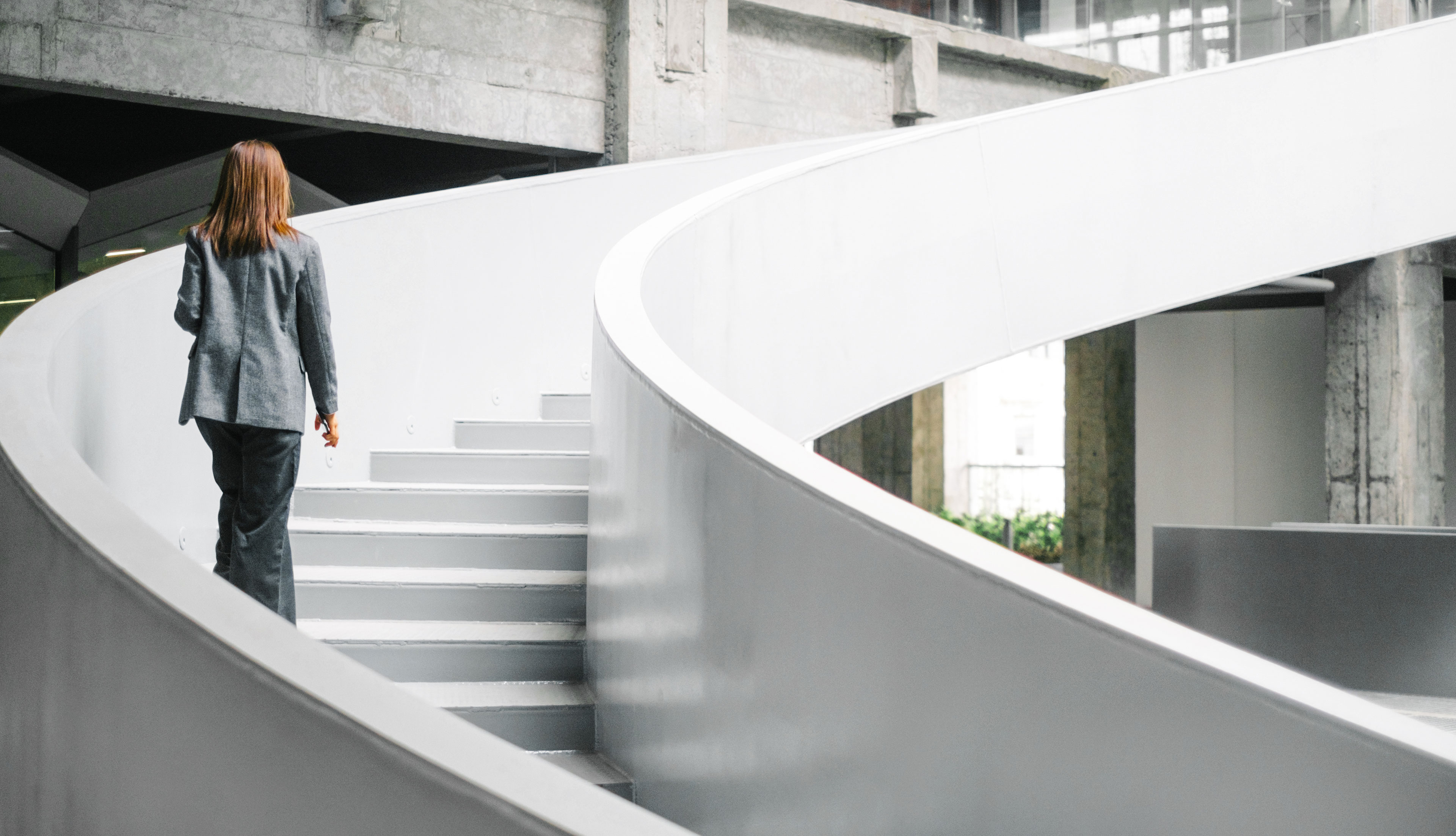 Femme d'affaires en costume gris montant des escaliers blancs en colimaçon vers l'étage suivant dans un immeuble de bureaux