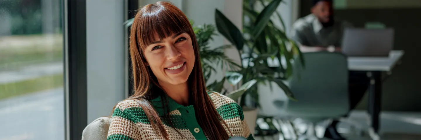 Une femme souriante aux cheveux foncés est assise confortablement dans un bureau lumineux et moderne et regarde chaleureusement la caméra. En arrière-plan, on distingue d’autres personnes et de grandes fenêtres.
