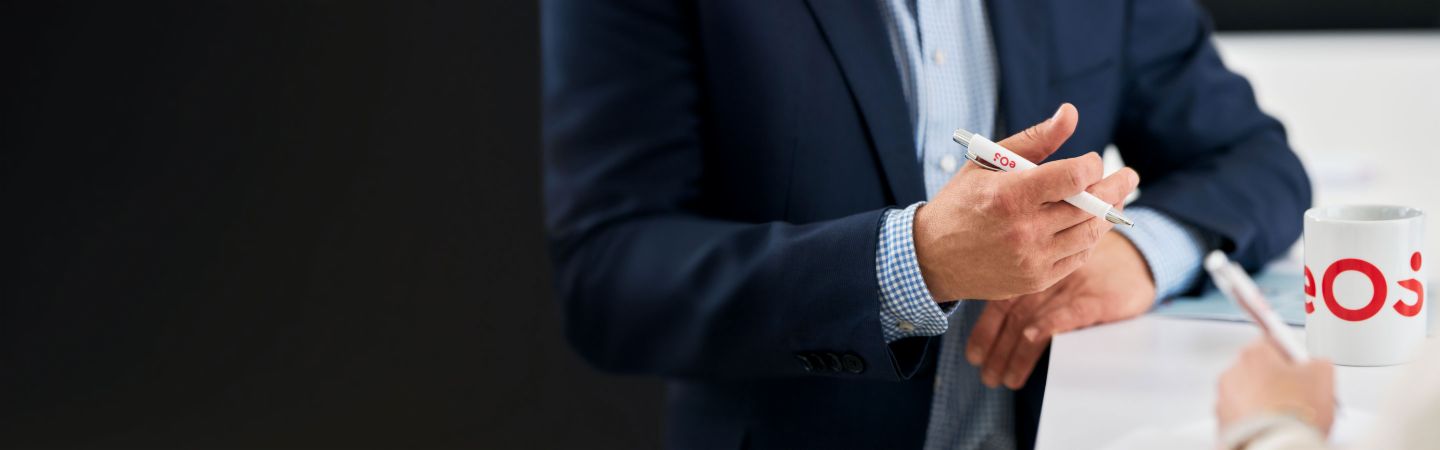 Homme d'affaires tenant un stylo EOS lors d'une conversation sur la gestion internationale des créances, tasse EOS sur la table - Expertise en recouvrement de créances, NPL et solutions numériques