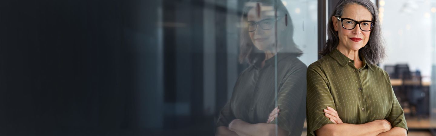L'image montre une femme debout avec confiance, les bras croisés, portant une chemise verte, devant un mur en verre qui reflète un espace de bureau moderne avec un éclairage lumineux et des meubles.