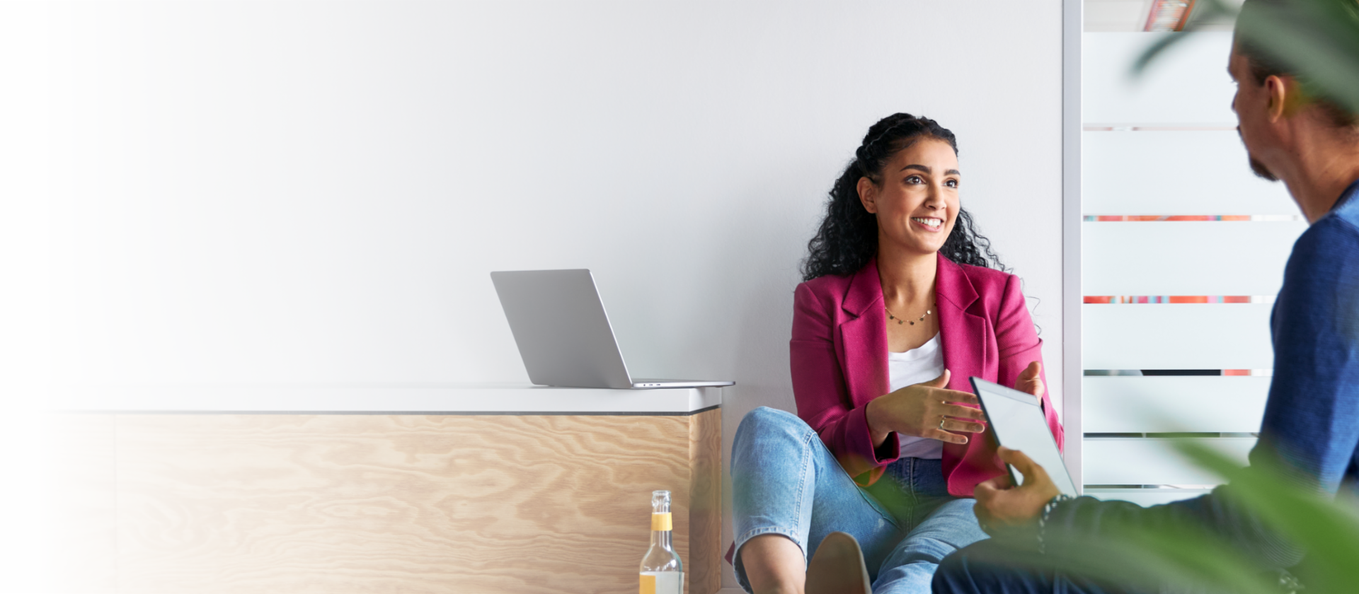 Deux jeunes femmes, celle de gauche portant un blazer rose et un pantalon noir et celle de droite portant une chemise blanche et un pantalon beige, se promènent côte à côte devant un mur blanc dans un bâtiment lumineux. Elles discutent. À l’avant, à droite, nous voyons une plante verte floue.