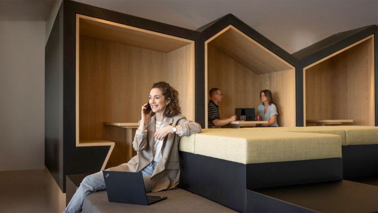 Une personne souriante est assise dans un salon de bureau moderne, parle au téléphone et travaille sur un ordinateur portable. En arrière-plan, deux autres personnes travaillent.
