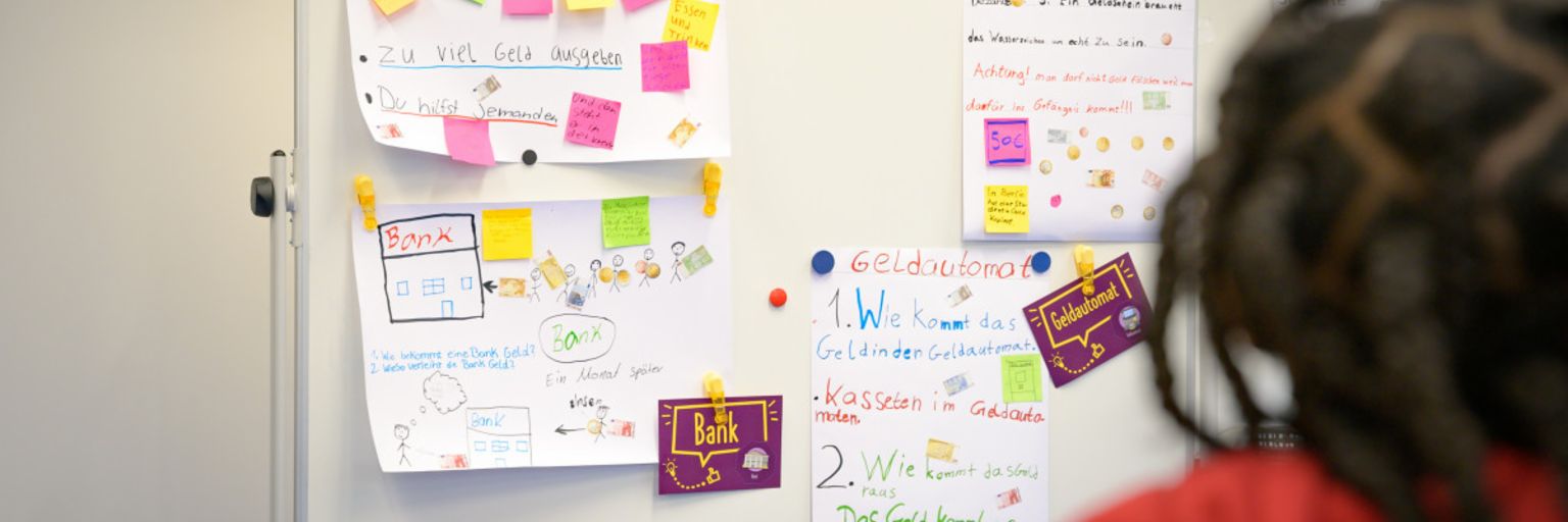 A photo shows post-its and paper notes on a board with terms like “Debt”, “Cash”, “Bank” and “ATM”. Underneath, the text reads: “In the ManoMoneta program developed by the EOS finlit foundation, schoolchildren aged 9 to 13 can learn about financial products.“