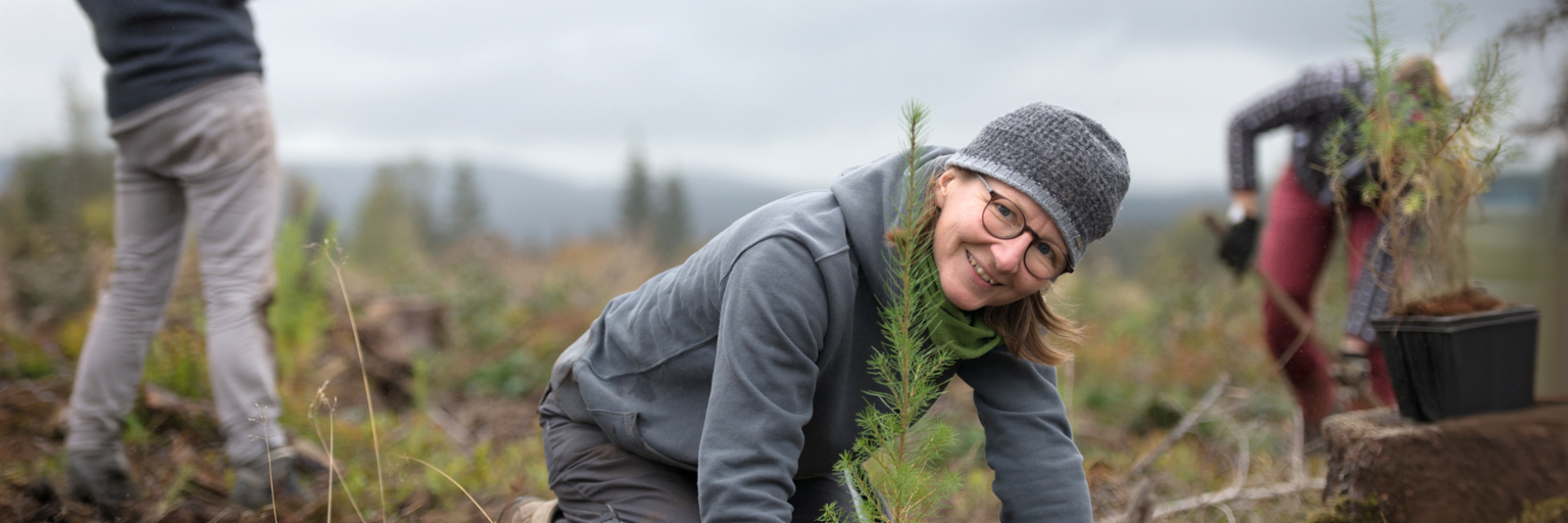 Une jeune femme portant un haut à capuche gris, un pantalon gris et une casquette grise, est agenouillée dans un champ devant un arbre qui vient d’être planté et elle sourit à la caméra. À l’arrière-plan, à droite, on discerne une autre personne portant un pantalon rouge qui creuse un trou pour planter un arbre. Dans le coin gauche, on devine l’image tronquée d’une personne qui tient un téléphone portable et s’apprête à prendre une photo.