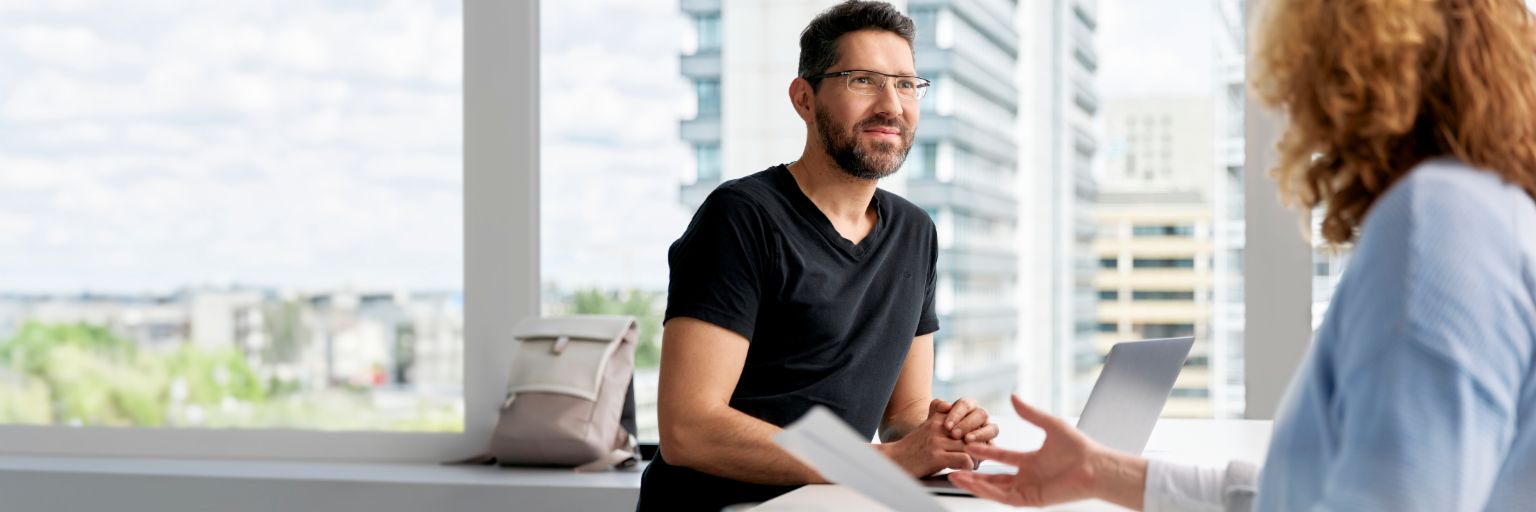 Deux personnes assises dans un bureau moderne et lumineux, en pleine conversation. <br/>Au centre de la photo, un homme à cheveux foncés, lunettes et barbe sourit. Il porte un T-shirt noir. Quelques documents et un ordinateur portable sont posés sur la table devant lui. La deuxième personne, dont nous voyons les bras et la tête, est assise en face de l’homme. Elle porte un chemisier léger, a des cheveux courts, bouclés et blond-roux et tient quelques feuilles de papier. À l’arrière-plan, on discerne de grandes fenêtres à travers lesquelles se dessinent des bâtiments modernes.