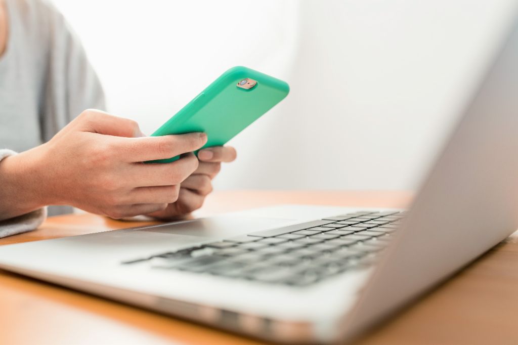 Woman holding smartphone while typing on laptop.