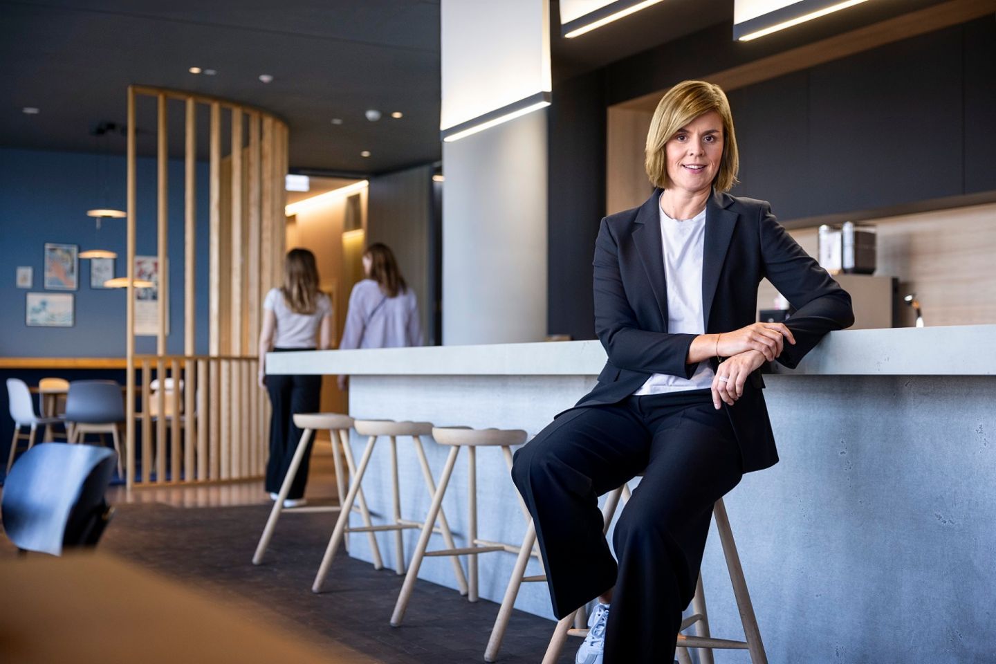 Dr. Eva Griewel, CFO du Groupe EOS, est assise dans un espace social sur un tabouret haut, portant un blazer noir.