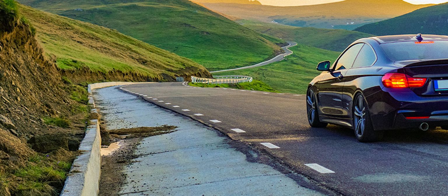 A photomontage shows a car traveling toward the sunset in the right lane of a paved road in an open, green landscape. The asphalt on the opposite lane is completely worn down.