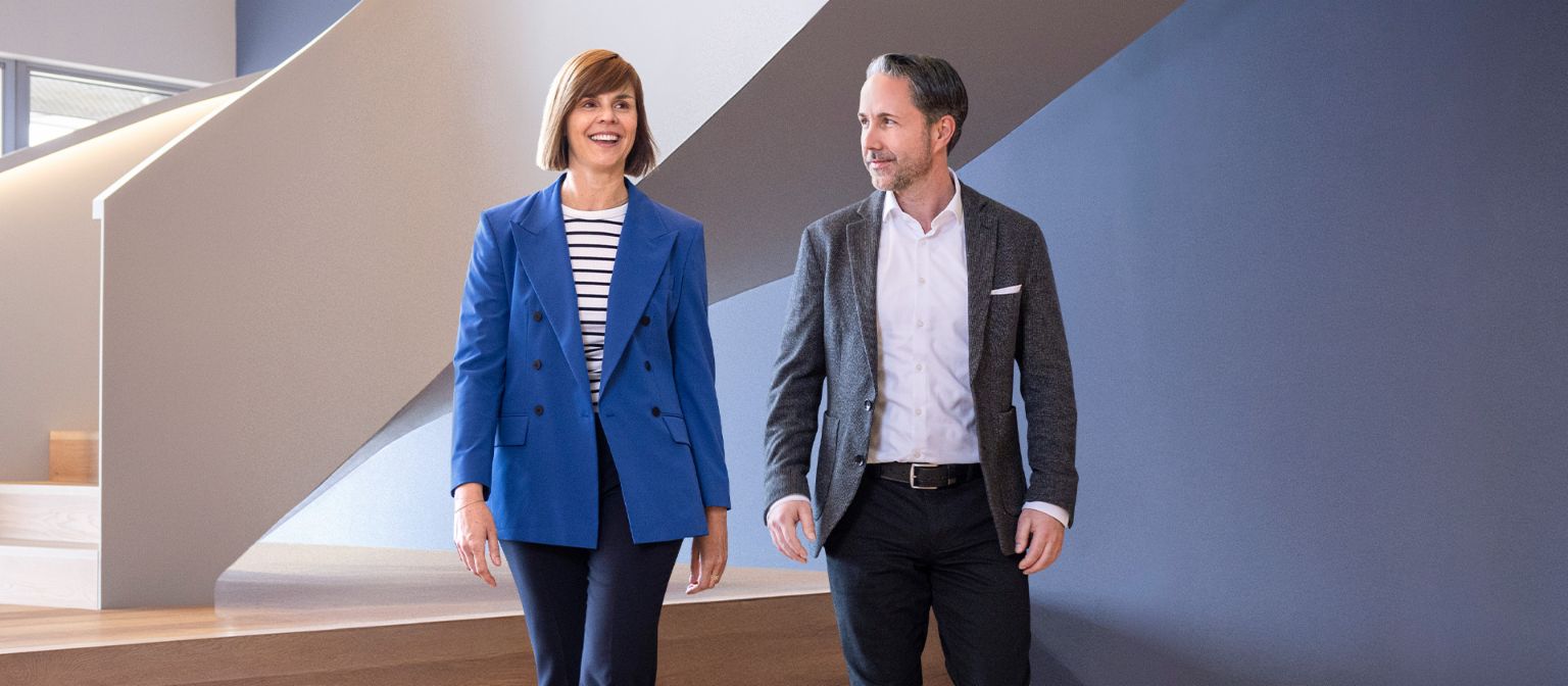 Le PDG en blazer gris et le CFO du Groupe EOS en blazer bleu traversent un bureau moderne. L'espace dispose de grandes fenêtres, d'un mur courbé et de lampes suspendues au design élégant.