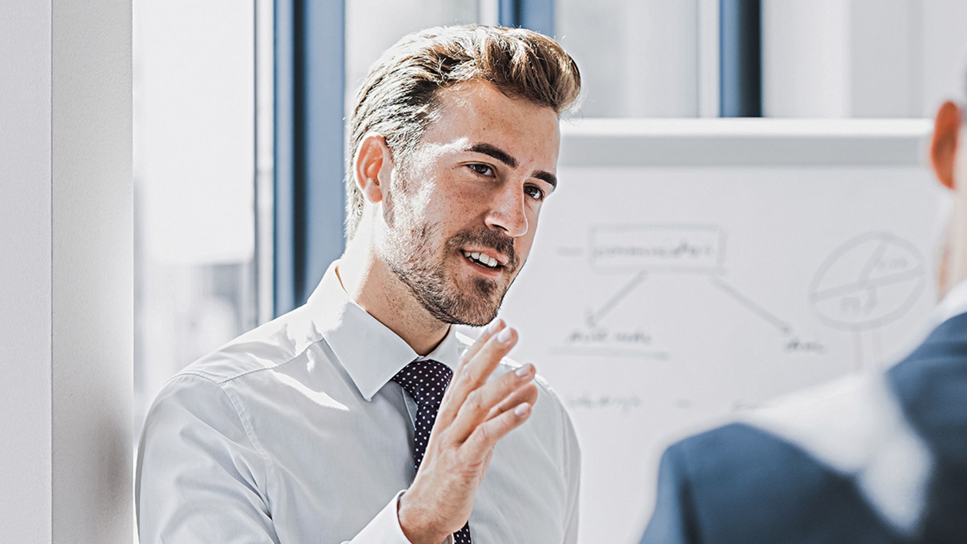 On voit un homme en tenue professionnelle expliquer quelque chose avec une expression amicale et engagée. Face à lui se tient une autre personne qui l’écoute attentivement. On aperçoit en arrière-plan un tableau blanc avec un croquis.