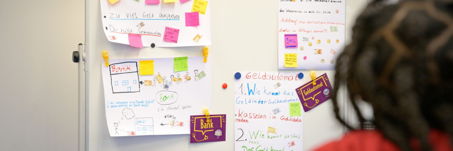 A photo shows post-its and paper notes on a board with terms like “Debt”, “Cash”, “Bank” and “ATM”. Underneath, the text reads: “In the ManoMoneta program developed by the EOS finlit foundation, schoolchildren aged 9 to 13 can learn about financial products.“