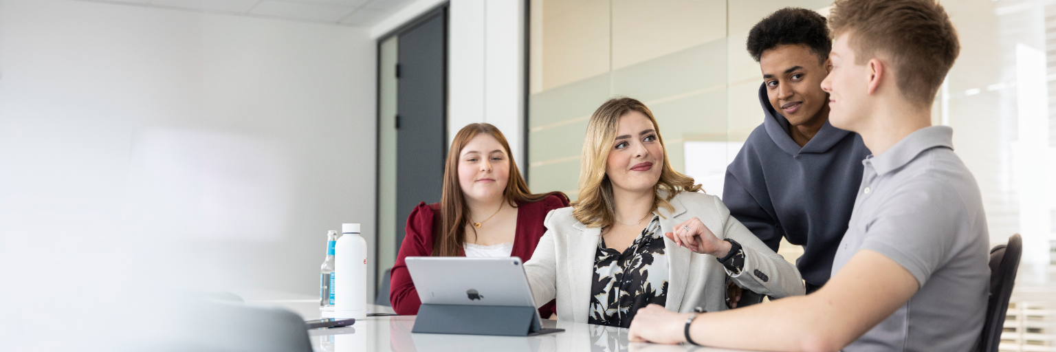 Quatre jeunes dans un bureau moderne, assis ou debout devant une table de conférence blanche. Une femme est assise au centre de l’image et regarde les autres avec un air bienveillant. Une tablette est posée sur la table devant elle. Deux autres personnes sont assises ou debout de part et d’autre d’elle. La personne de gauche porte une veste rouge et esquisse un léger sourire. L’autre personne porte un haut à capuche gris et regarde l’homme portant un polo clair à droite de la photo, qui regarde la femme à la tablette.