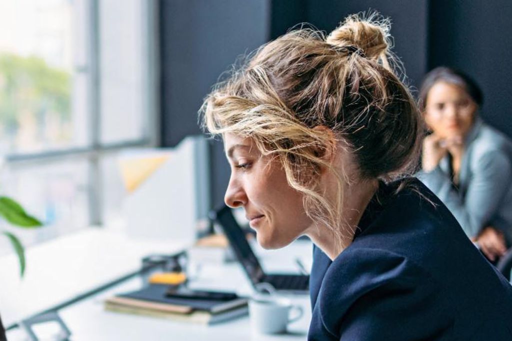 L'image montre une femme assise à un bureau dans un bureau moderne, portant un blazer sombre tout en se concentrant sur son ordinateur portable, avec une plante et quelques fournitures de bureau visibles sur la table, et une autre personne assise en arrière-plan.